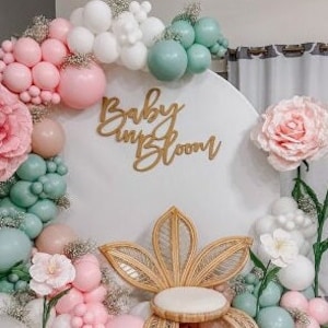 May include: A white backdrop with a round arch decorated with pink, white, and green balloons, paper flowers, and greenery. The text "Baby in Bloom" is displayed in gold lettering on the backdrop. A wicker chair with a white cushion is in the foreground.