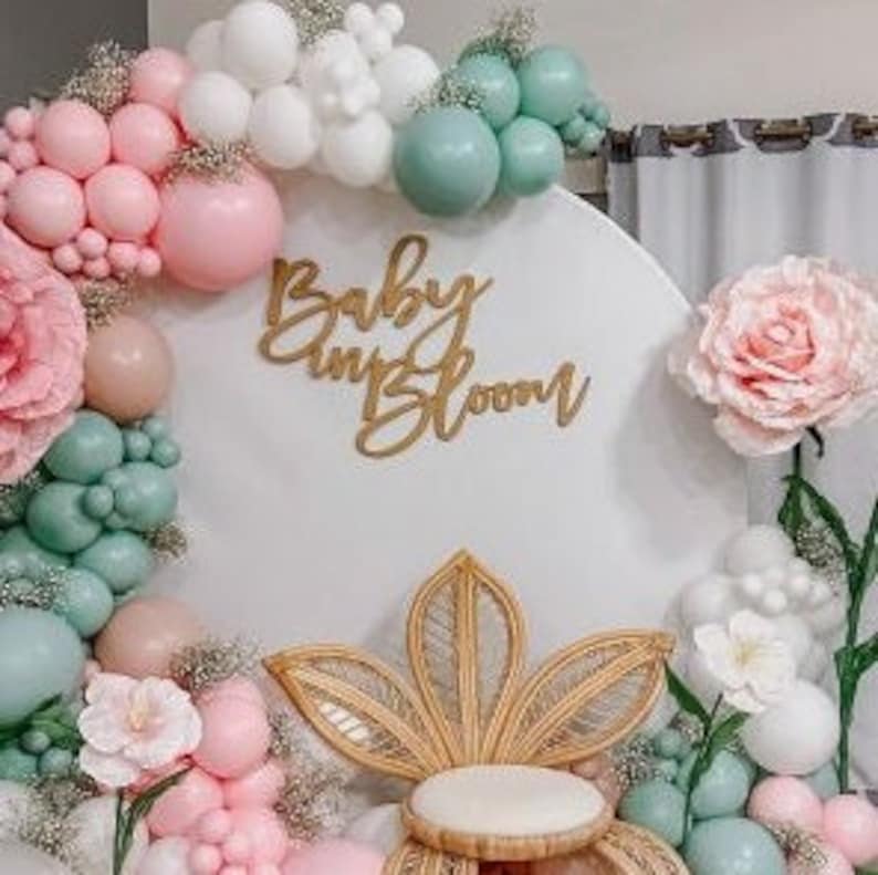 May include: A white backdrop with a round arch decorated with pink, white, and green balloons, paper flowers, and greenery. The text "Baby in Bloom" is displayed in gold lettering on the backdrop. A wicker chair with a white cushion is in the foreground.