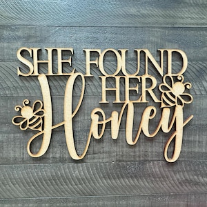 May include: Wooden sign with the phrase "SHE FOUND HER HONEY" in a decorative font. The word "Honey" is in cursive, with bee illustrations on either side. The sign is made of light-colored wood and is set against a gray wood-grain background.