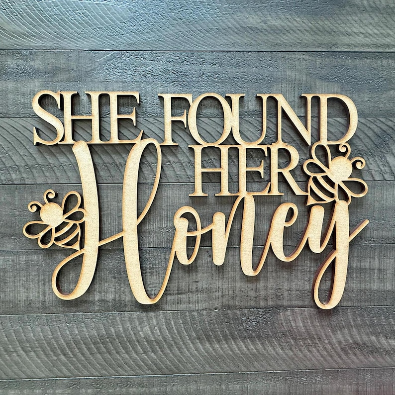 May include: Wooden sign with the phrase "SHE FOUND HER HONEY" in a decorative font. The word "Honey" is in cursive, with bee illustrations on either side. The sign is made of light-colored wood and is set against a gray wood-grain background.