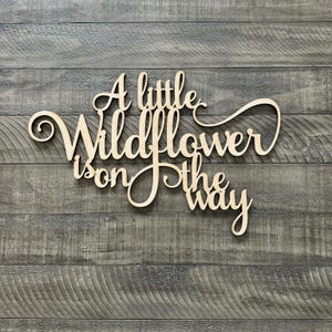 以下が含まれることがあります： 「A little Wildflower is on the way」という文字が書かれた木製の看板。筆記体で書かれています。