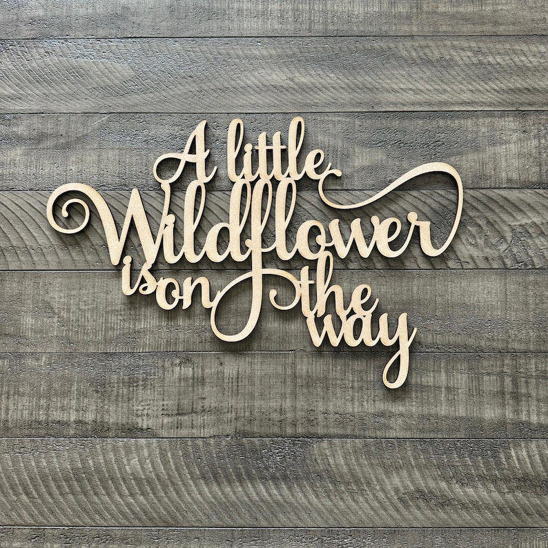 以下が含まれることがあります： 「A little Wildflower is on the way」という文字が書かれた木製の看板。筆記体で書かれています。