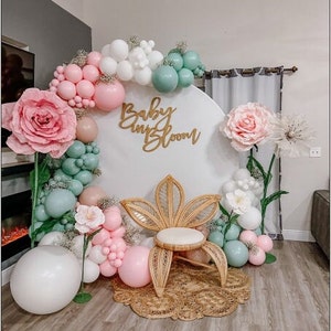 Op de afbeelding: Een witte achtergrond met een gouden bord waarop 'Baby in Bloom' staat, omringd door roze en groene ballonnen, witte papieren bloemen en groen. Een rieten stoel met een wit kussen staat voor de achtergrond.