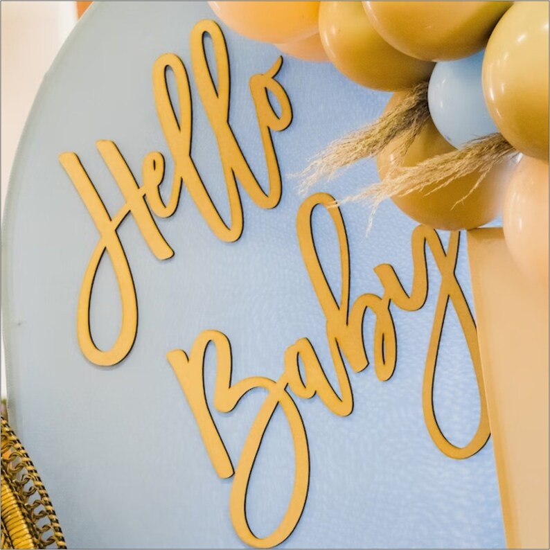 May include: Gold cursive lettering on a light blue background that reads "Hello Baby".