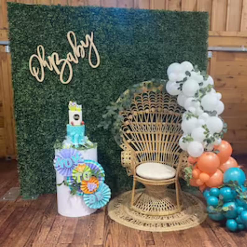 May include: A baby shower backdrop with a green artificial grass wall, a wooden sign that says "Oh Baby", a white wicker peacock chair, and a white cake stand with a blue and white cake. There are also blue, green, and orange balloons.