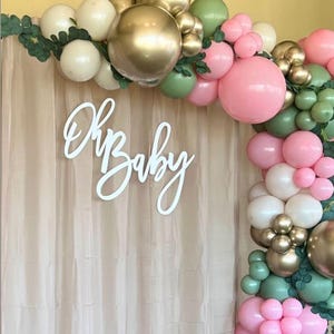 May include: A white wooden sign with the text "Oh Baby" in cursive script, hanging in front of a beige backdrop. The sign is decorated with a garland of green, pink, and cream balloons.