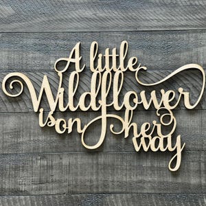 以下が含まれることがあります： 「A little Wildflower is on her way」という文字が書かれた木製の看板。筆記体で書かれています。
