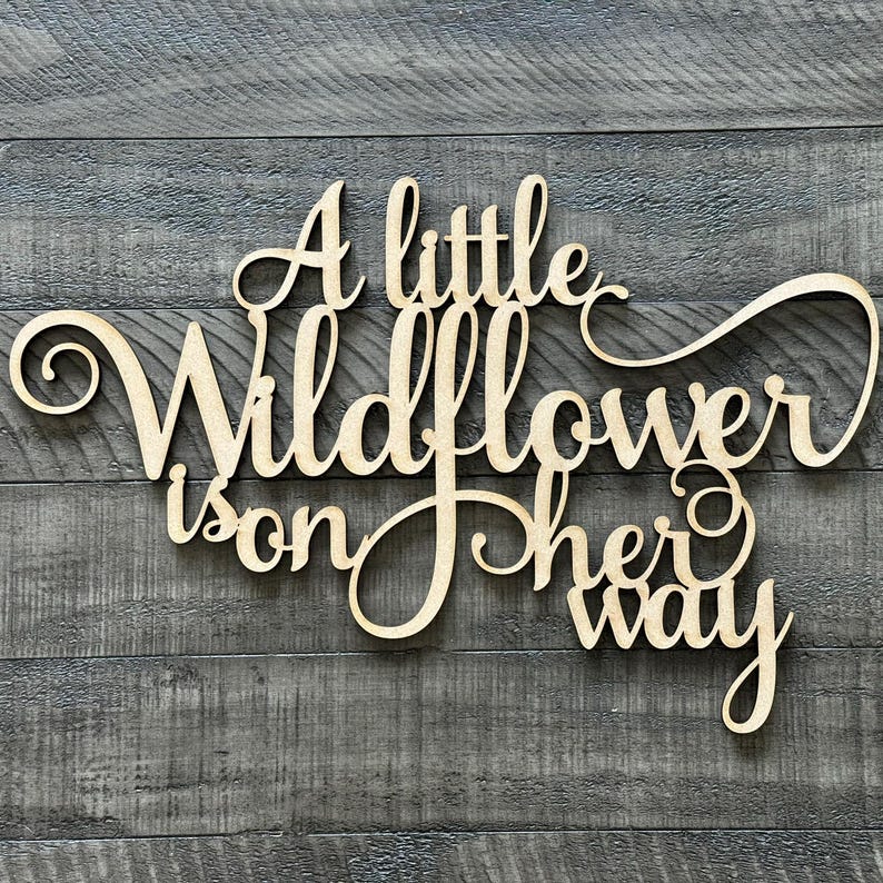 以下が含まれることがあります： 「A little Wildflower is on her way」という文字が書かれた木製の看板。筆記体で書かれています。