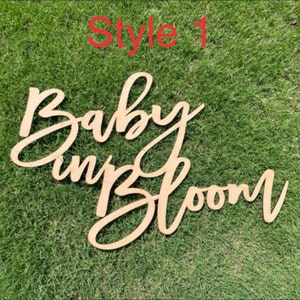 May include: Wooden sign with the text "Baby in Bloom" in a cursive font. The sign is light brown and is on a green grassy background.