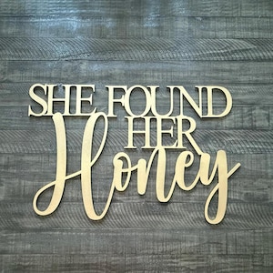 May include: Wooden sign with the phrase "SHE FOUND HER Honey" in a mix of serif and cursive fonts. The sign is light brown and mounted on a gray wooden plank background. The sign is a decorative item.