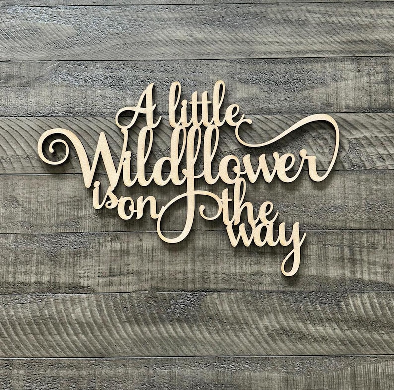 以下が含まれることがあります： 「A little Wildflower is on the way」という文字が書かれた木製の看板。筆記体で書かれています。