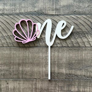 May include: A white wooden cake topper with the word "one" in a cursive font. A pink seashell design is on the left side of the word. The topper is designed to be inserted into a cake. The background is a wooden surface.
