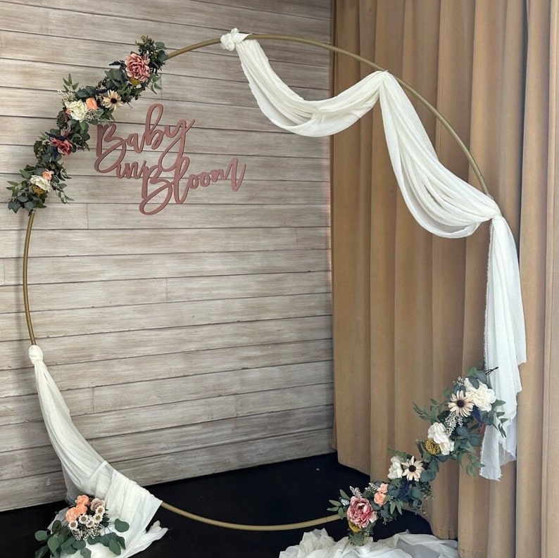 May include: A gold metal hoop arch decorated with white fabric and pink and white flowers. The arch is set against a wood panel wall with the words "Baby in Bloom" written in pink.