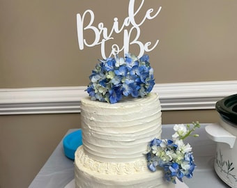 Op de afbeelding: Een witte bruidstaart met twee lagen, versierd met blauwe hortensia's en een witte 'Bride to Be'-taarttopper.