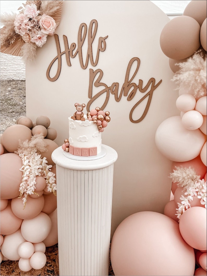 May include: A white cake stand with a small white cake decorated with a teddy bear and pink and gold balloons. The cake stand is on a white pedestal with a wood grain texture. The background is a white wall with a wooden sign that says "Hello Baby".