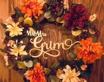 May include: A fall-themed wreath with orange, yellow, and burgundy flowers, greenery, and a wooden sign that reads "Mr. & Mrs. Grim".