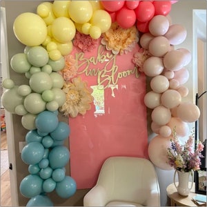 Op de afbeelding: Een roze muur versierd met een ballonboog in tinten geel, groen, blauw en roze. De boog is gemaakt van ballonnen in verschillende maten. De muur heeft ook een houten bord met de tekst "Baby in Bloom".