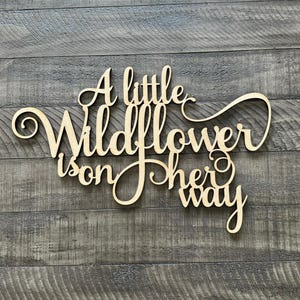 以下が含まれることがあります： 「A little wildflower is on her way」と書かれた木製の看板。看板は薄い茶色の木でできており、素朴な外観をしています。