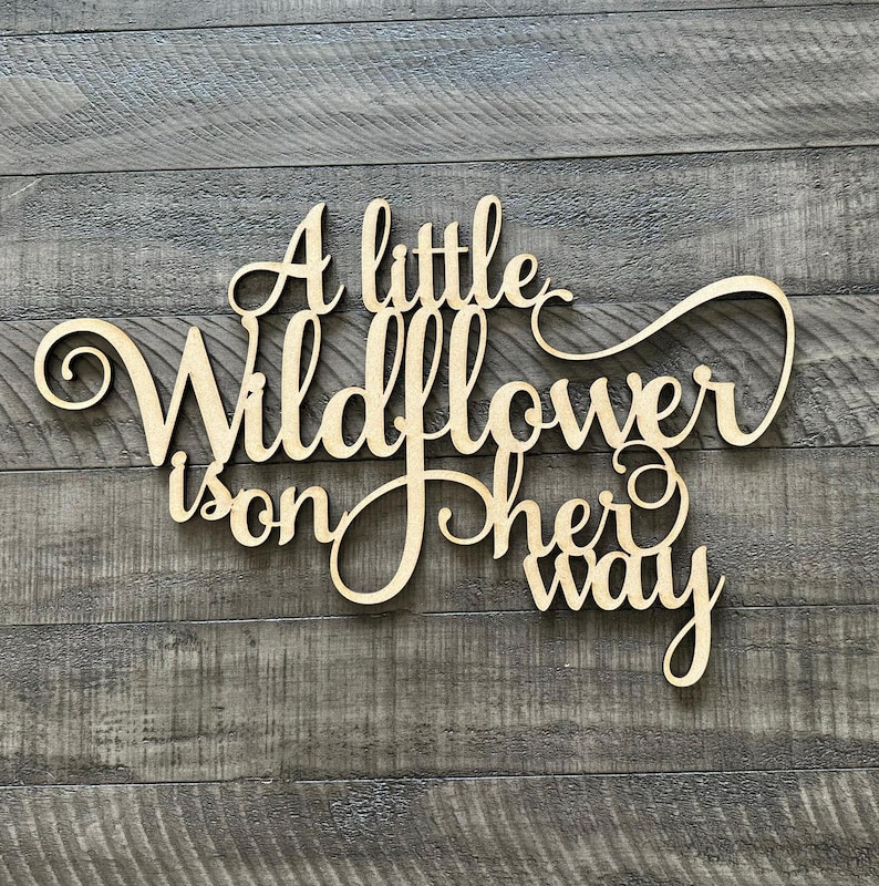 以下が含まれることがあります： 「A little wildflower is on her way」と書かれた木製の看板。看板は薄い茶色の木でできており、素朴な外観をしています。