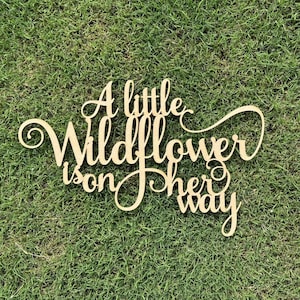 以下が含まれることがあります： 「A little wildflower is on her way」と書かれた木製の看板。看板は薄い茶色に塗られており、素朴でヴィンテージな雰囲気です。