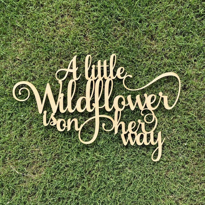 以下が含まれることがあります： 「A little wildflower is on her way」と書かれた木製の看板。看板は薄い茶色に塗られており、素朴でヴィンテージな雰囲気です。
