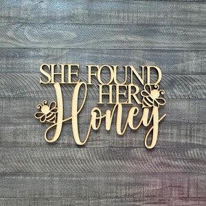 May include: Wooden sign with the phrase "SHE FOUND HER Honey" in a decorative font. The word "Honey" is in cursive, with bee illustrations on either side. The sign is made of light-colored wood and is set against a gray wood-grain background.