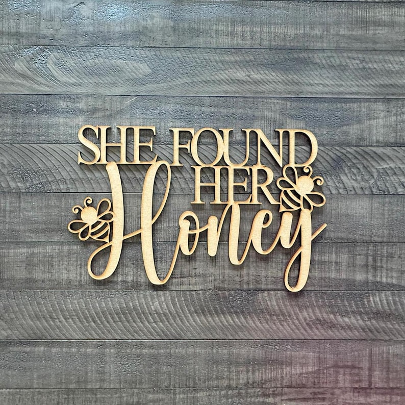 May include: Wooden sign with the phrase "SHE FOUND HER Honey" in a decorative font. The word "Honey" is in cursive, with bee illustrations on either side. The sign is made of light-colored wood and is set against a gray wood-grain background.