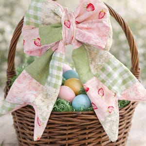 Może przedstawiać: Brązowy wiklinowy kosz wielkanocny ozdobiony dużą patchworkową kokardą z truskawkami, kratką i kwiatowymi wzorami. Kosz zawiera kolorowe jajka osadzone w zielonej trawie dekoracyjnej, tworząc świąteczną wiosenną scenę.