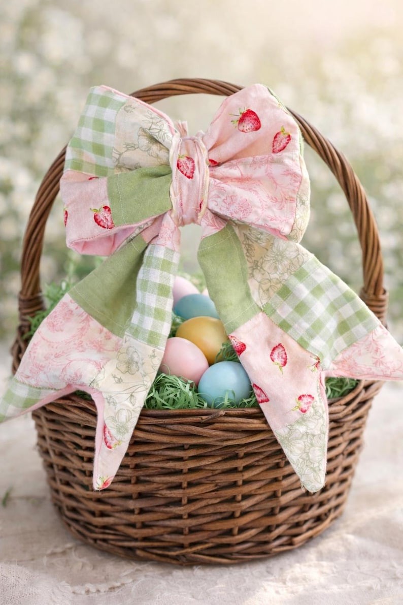 Może przedstawiać: Brązowy wiklinowy kosz wielkanocny ozdobiony dużą patchworkową kokardą z truskawkami, kratką i kwiatowymi wzorami. Kosz zawiera kolorowe jajka osadzone w zielonej trawie dekoracyjnej, tworząc świąteczną wiosenną scenę.