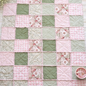 May include: A patchwork quilt in a grid pattern, with pink, green, and floral fabric squares. The quilt has a ruffled edge and is on a light wood surface. A small bowl of buttons is in the bottom right corner.