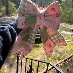 Może przedstawiać: Duża, ręcznie robiona kokarda z tkaniny o wzorze patchworku. Kokarda zawiera r&oacute;żowy materiał z wzorami truskawek, zieloną kratę vichy i nadrukami kwiatowymi. Kokarda jest prezentowana na naturalnym tle zewnętrznym.