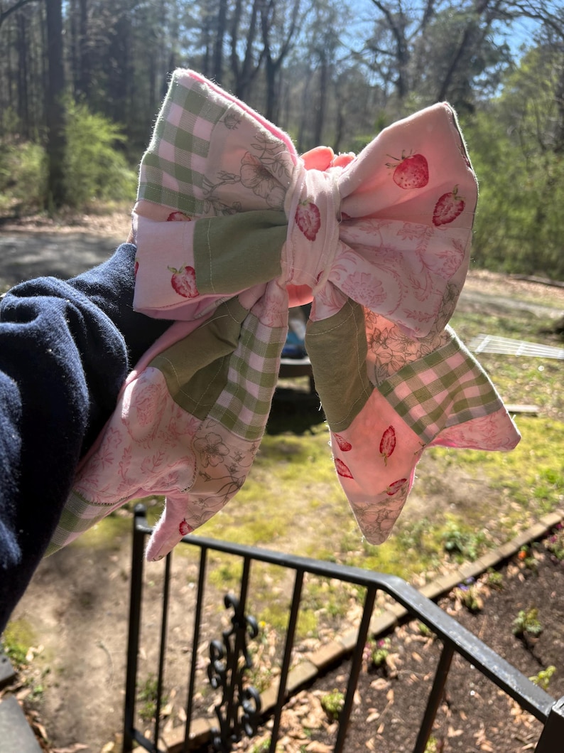 Może przedstawiać: Duża, ręcznie robiona kokarda z tkaniny o wzorze patchworku. Kokarda zawiera r&oacute;żowy materiał z wzorami truskawek, zieloną kratę vichy i nadrukami kwiatowymi. Kokarda jest prezentowana na naturalnym tle zewnętrznym.