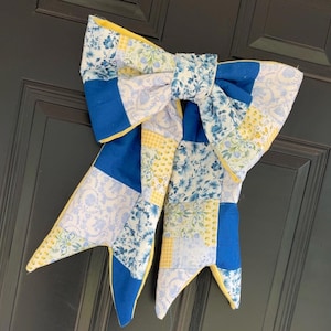 May include: A large, decorative bow made of patchwork fabric. The bow features a combination of blue floral, yellow gingham, and solid blue fabric pieces. Yellow fabric edges the bow's design, creating a striking contrast against the dark background.