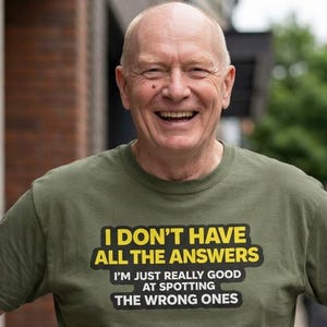 May include: An olive green short-sleeved t-shirt featuring the text "I DON'T HAVE ALL THE ANSWERS I'M JUST REALLY GOOD AT SPOTTING THE WRONG ONES" printed in yellow and white with black outlines on the chest area of the garment.