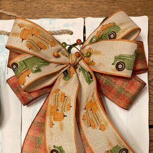 Könnte beinhalten: Eine dekorative Schleife mit einem beigen Band, bedruckt mit orangefarbenen Kürbissen und grünen Lastwagen. Die Schleife hat eine karierte orange-rote Bandbasis. Die Schleife ist mit einem kleinen Zweig Beeren gebunden.