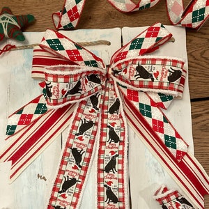 May include: A festive Christmas bow with red, white, and green ribbons. The bow features a plaid pattern with black dog silhouettes wearing Santa hats and the text "Merry Christmas, We Woof You". Additional star-shaped and striped decorations are also present.