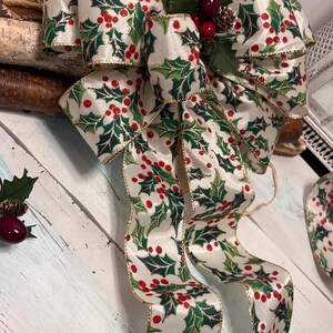 以下が含まれることがあります： 白いリボンで作られたクリスマスの装飾用リボン。ヒイラギの葉と赤い実の模様があり、金色のトリムが付いています。リボンは長く流れるようなテールで結ばれています。