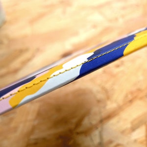 May include: Close-up of a blue, yellow, and pink patterned strap with yellow stitching. The strap is made of a soft, flexible material.