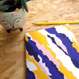 May include: A notebook with a colorful abstract design featuring yellow, pink, white, and blue swirls. The notebook is lying on a wooden surface.
