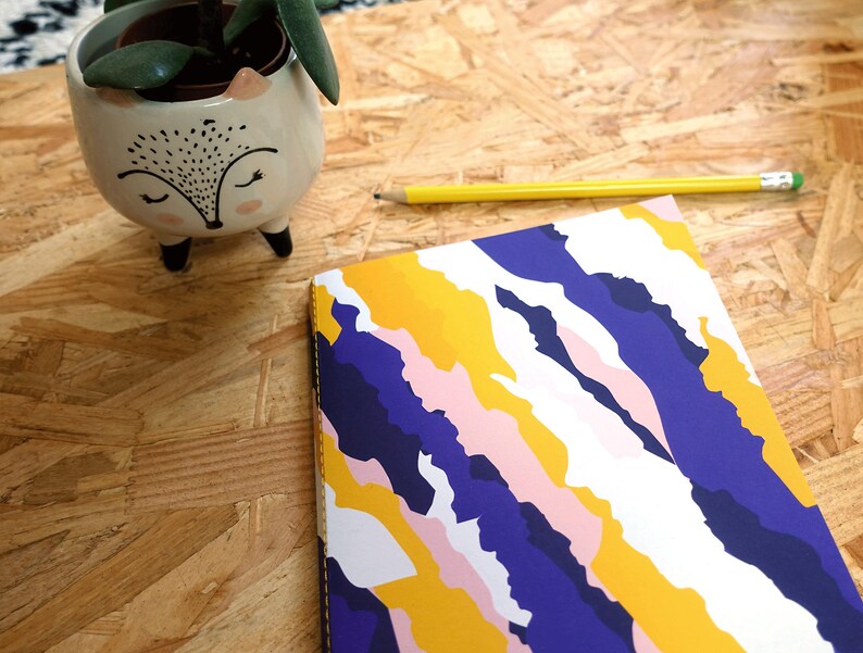 May include: A notebook with a colorful abstract design featuring yellow, pink, white, and blue swirls. The notebook is lying on a wooden surface.