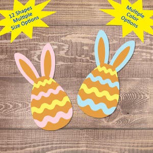 May include: Two Easter eggs with bunny ears. One egg is light blue with yellow wavy stripes and the other is pink with yellow wavy stripes. Both eggs are on a brown wooden background.