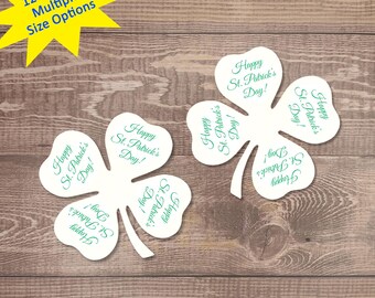 以下が含まれることがあります： 白い紙でできたクローバーの形が12個あり、それぞれに「Happy St. Patrick's Day!」という文字が印刷されています。クローバーは木目調の背景に集まって配置されています。