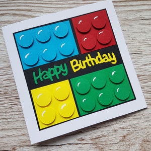 May include: A handmade birthday card featuring a colourful Lego block design with the words "Happy Birthday" in yellow lettering.