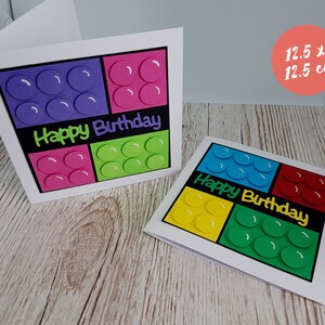 May include: Two handmade birthday cards featuring a colourful Lego block design. The cards are 12.5 cm by 12.5 cm and feature the words "Happy Birthday" in black.