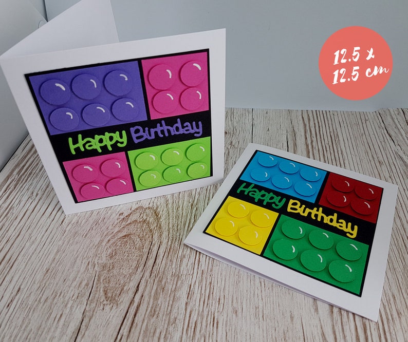 May include: Two handmade birthday cards featuring a colourful Lego block design. The cards are 12.5 cm by 12.5 cm and feature the words "Happy Birthday" in black.