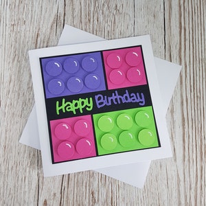 May include: A handmade birthday card with a Lego theme. The card features four square blocks with circles in purple, pink, and green. The text "Happy Birthday" is written in black on a black background.