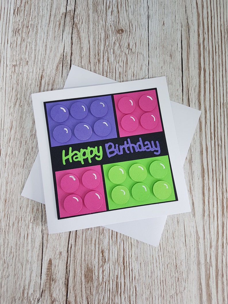 May include: A handmade birthday card with a Lego theme. The card features four square blocks with circles in purple, pink, and green. The text "Happy Birthday" is written in black on a black background.