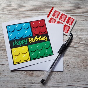 May include: A white card with a happy birthday message and a Lego-themed design. The card features four squares of different coloured Lego bricks, each with a white outline. The card is on a wooden surface with a pen and a sheet of stamps.