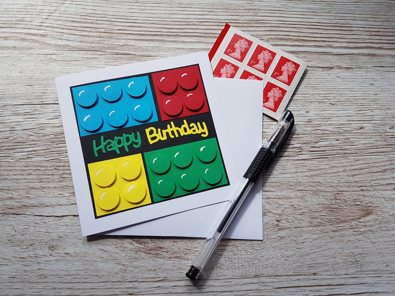 May include: A white card with a happy birthday message and a Lego-themed design. The card features four squares of different coloured Lego bricks, each with a white outline. The card is on a wooden surface with a pen and a sheet of stamps.