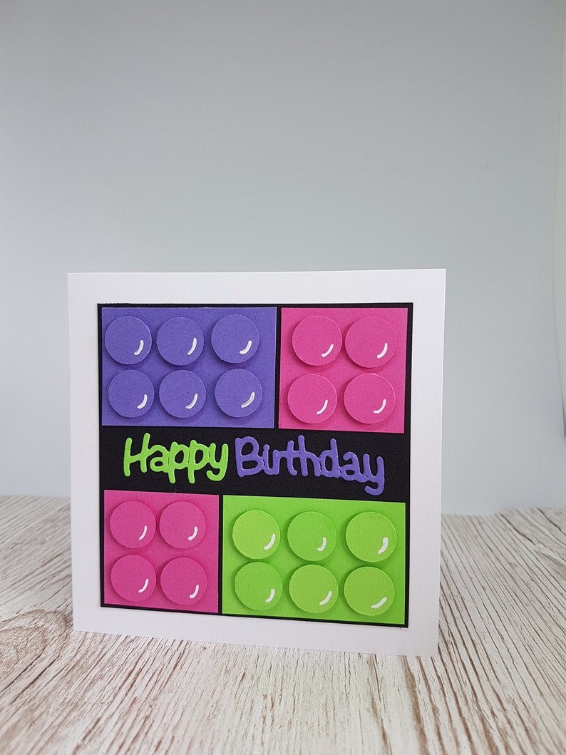 May include: A white card with a black background and the words "Happy Birthday" in green and purple lettering. The card is decorated with four colourful squares, each with a pattern of circles.